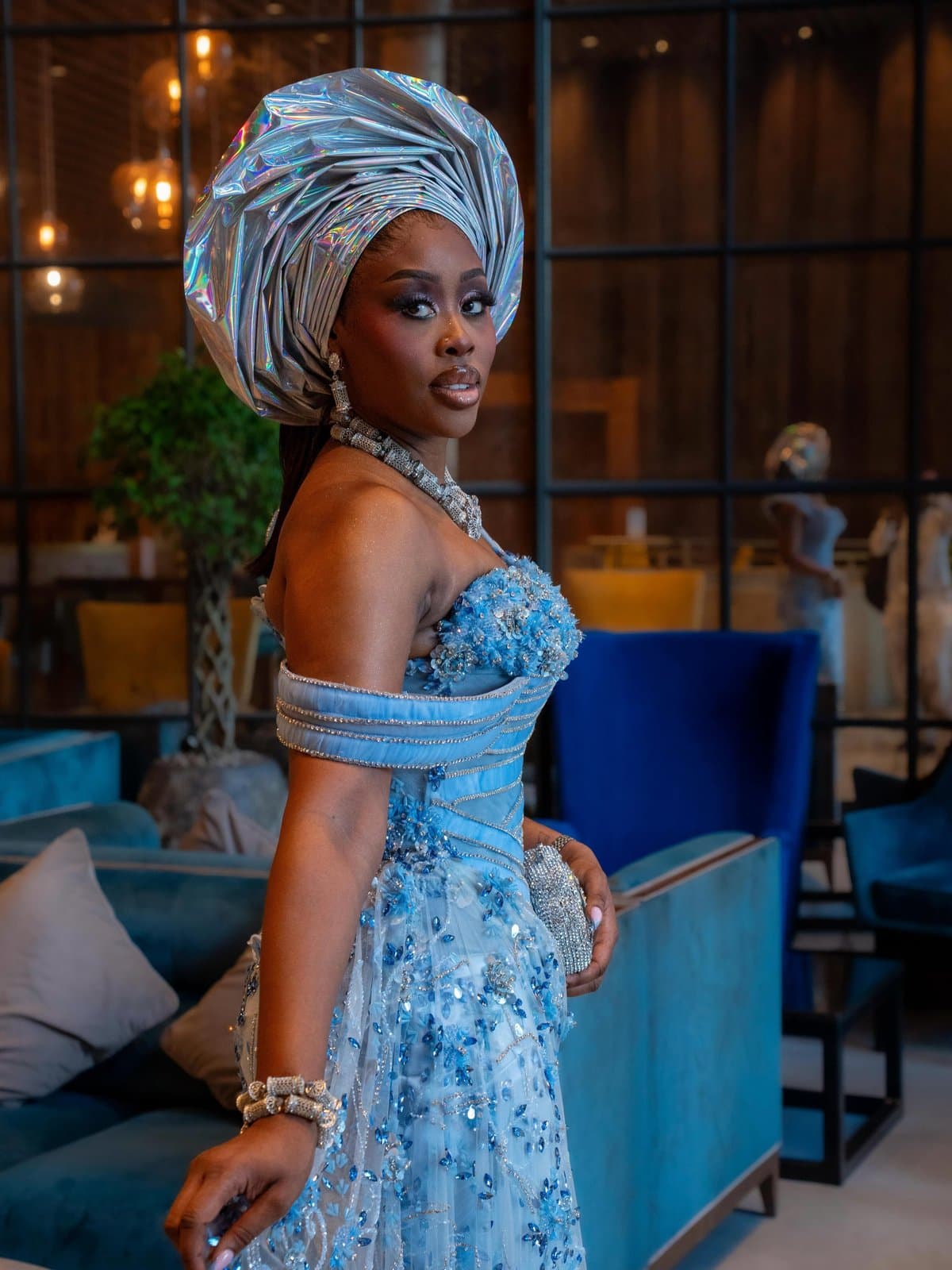 Silver gele catching every light in the room, profile turned toward the glow.